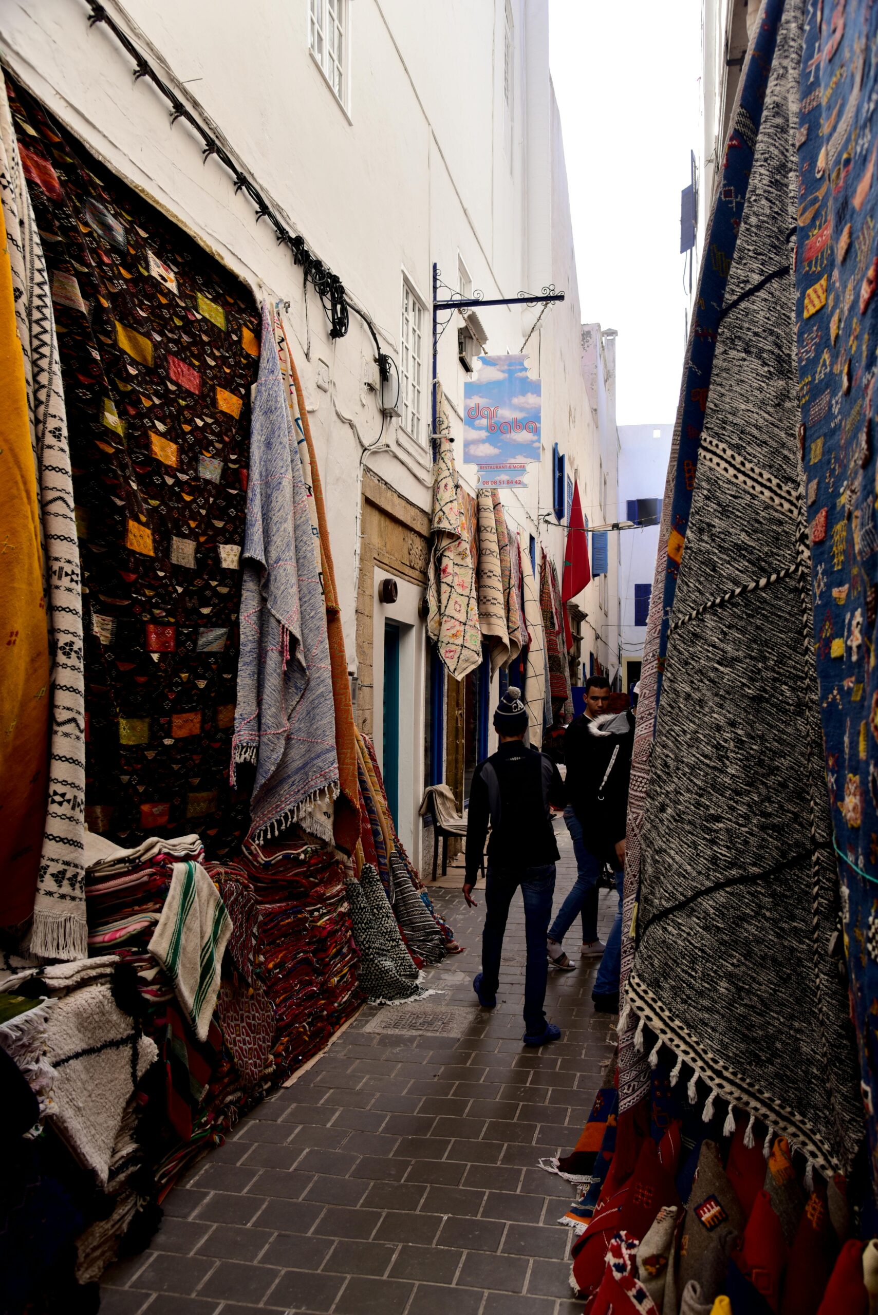 Colorful carpets displayed in a vibrant alley in Essaouira, Morocco with people exploring the souk.