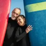 Smiling couple holding surfboards at a sunny beach, enjoying leisure time.
